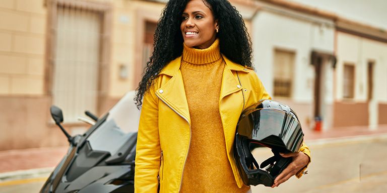 Mulher de casaco e blusa amarela, com capacete debaixo do braço e mota ao lado