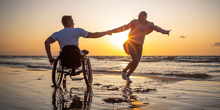 Homem em cadeiras de roda e mulher a dançar ao por do sol