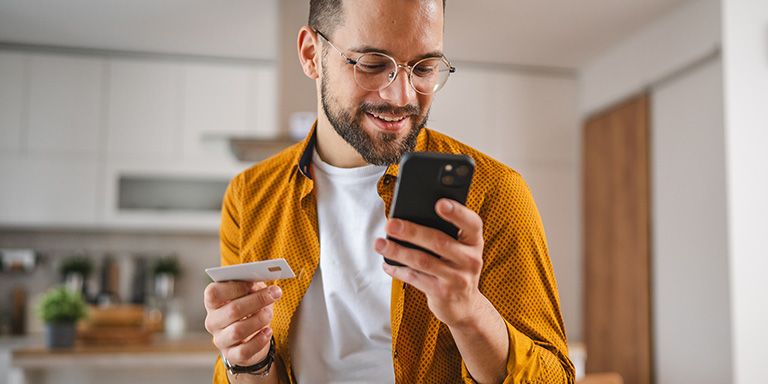 Homem de barba e óculos, camisola amarelo torrado, olha para o telemóvel para tentar perceber como poupar nas compras online