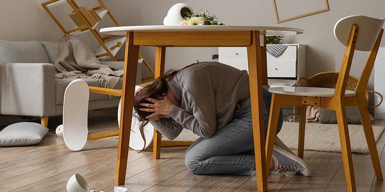 Rapariga ajoelha-se debaixo da mesa para se proteger de terramoto. Já pensou em adicionar a cobertura de sismos no seu seguro?